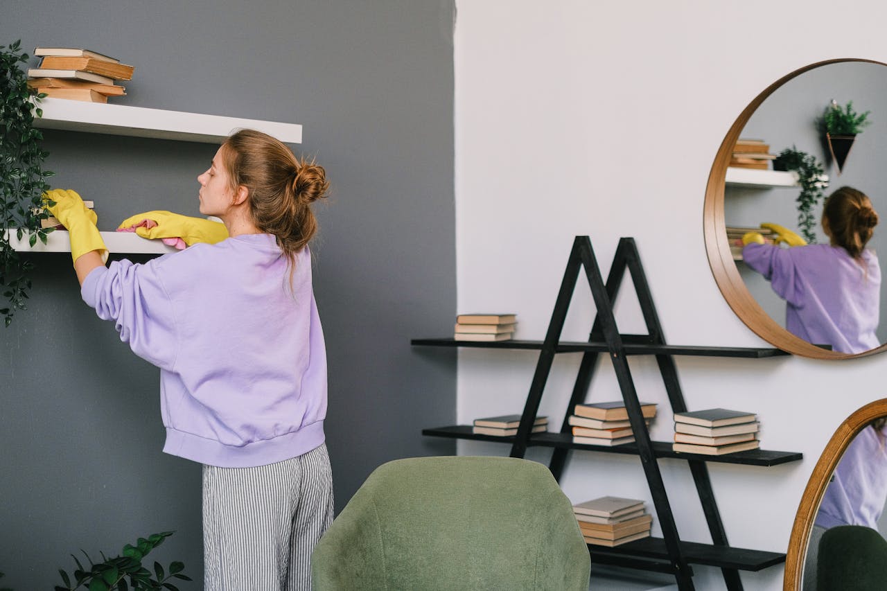 A woman cleans shelves in a stylish living room with mirrors and decor.