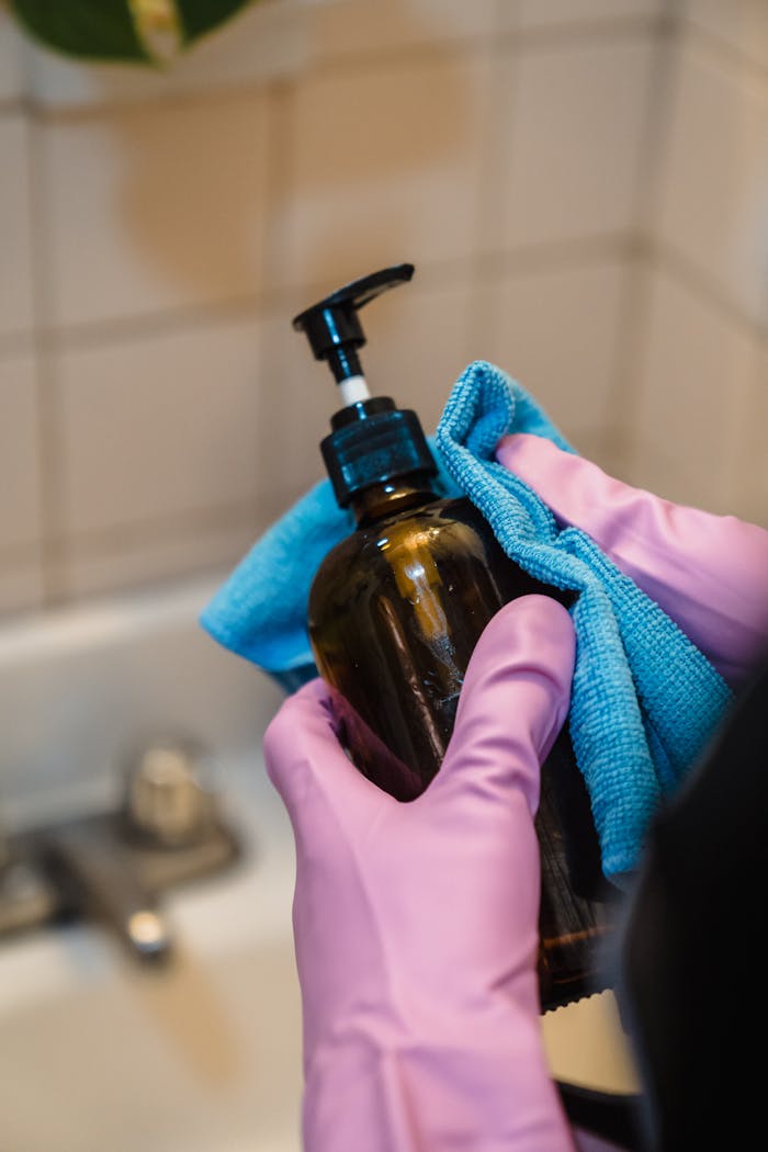 Close-up of hands cleaning a soap dispenser in a bathroom with blue cloth and latex gloves.